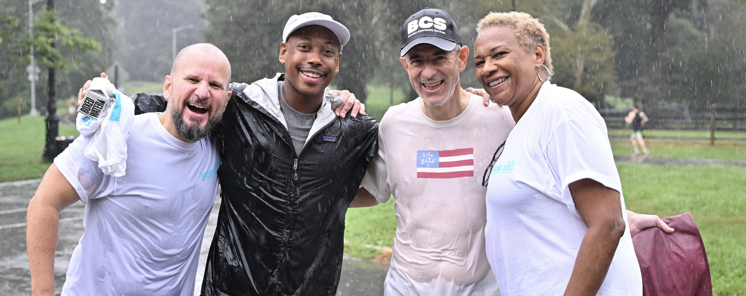 Four people with their arms around each other smile in the pouring rain.