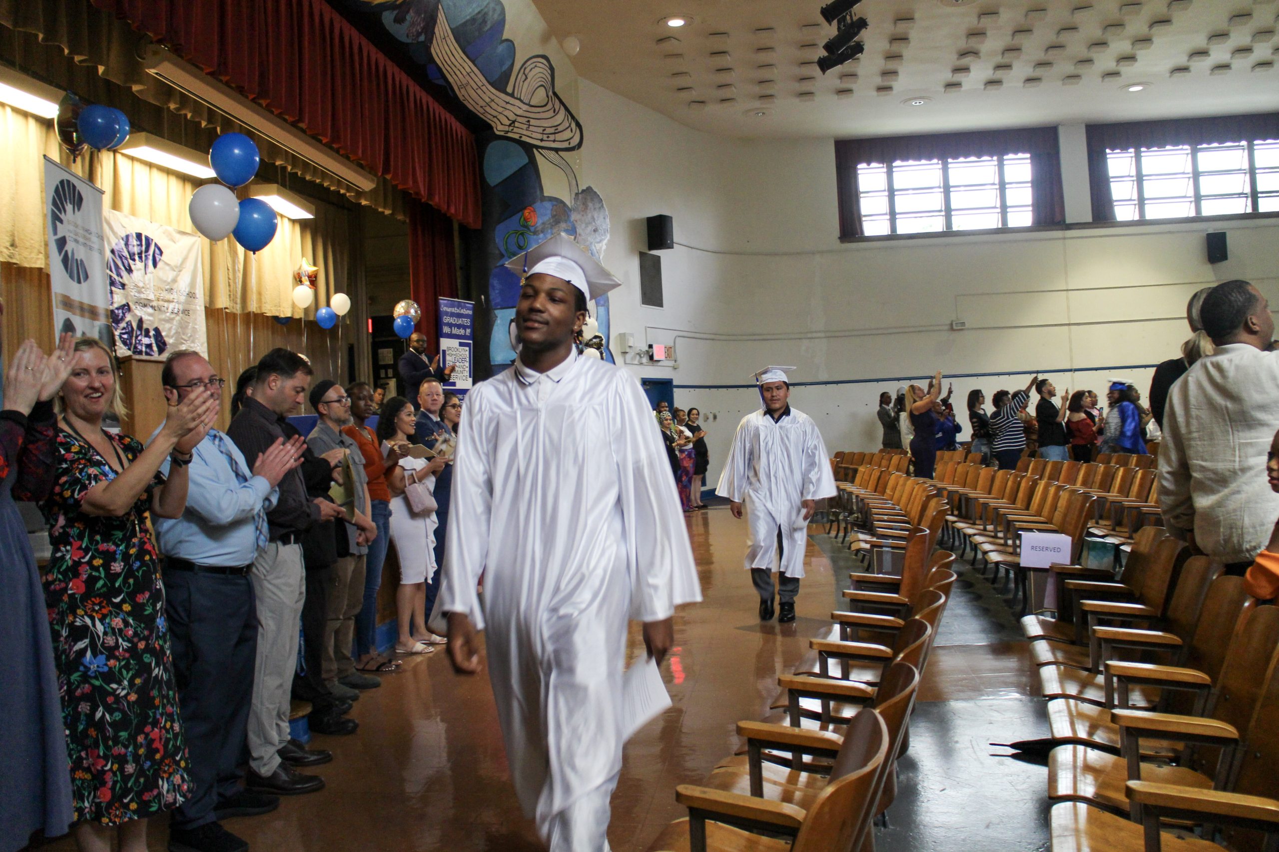 Inside a school auditorium, students in white caps and gowns walk past a line of teachers clapping.